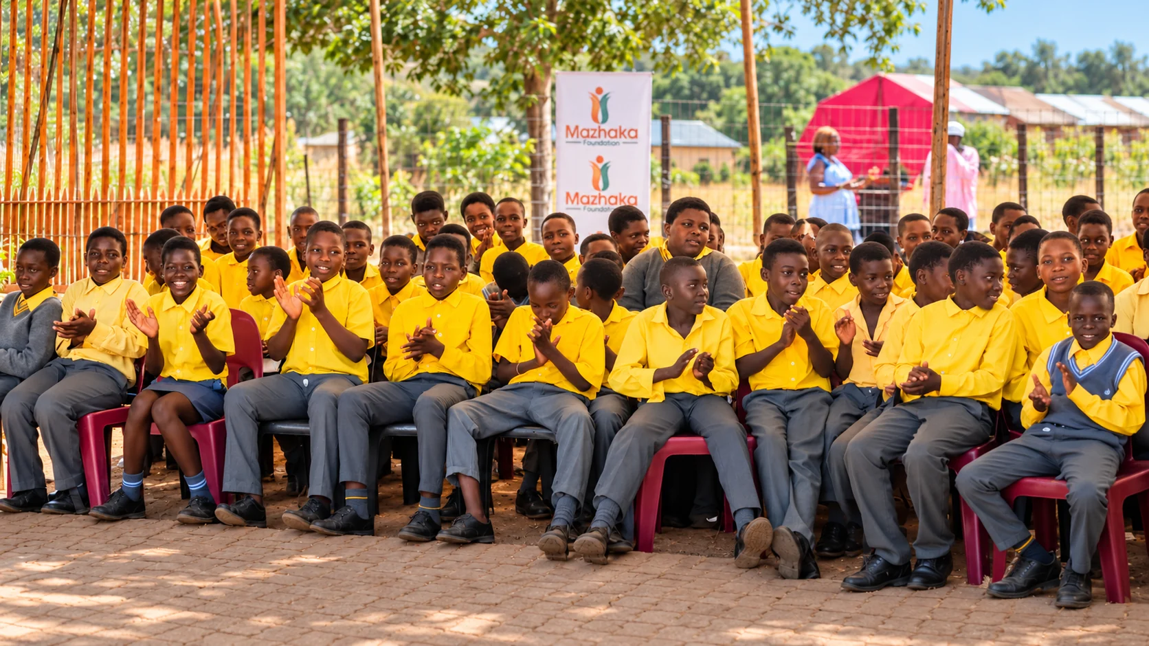 Schoolchildren in yellow uniforms applaud beneath a Mazhaka Foundation banner