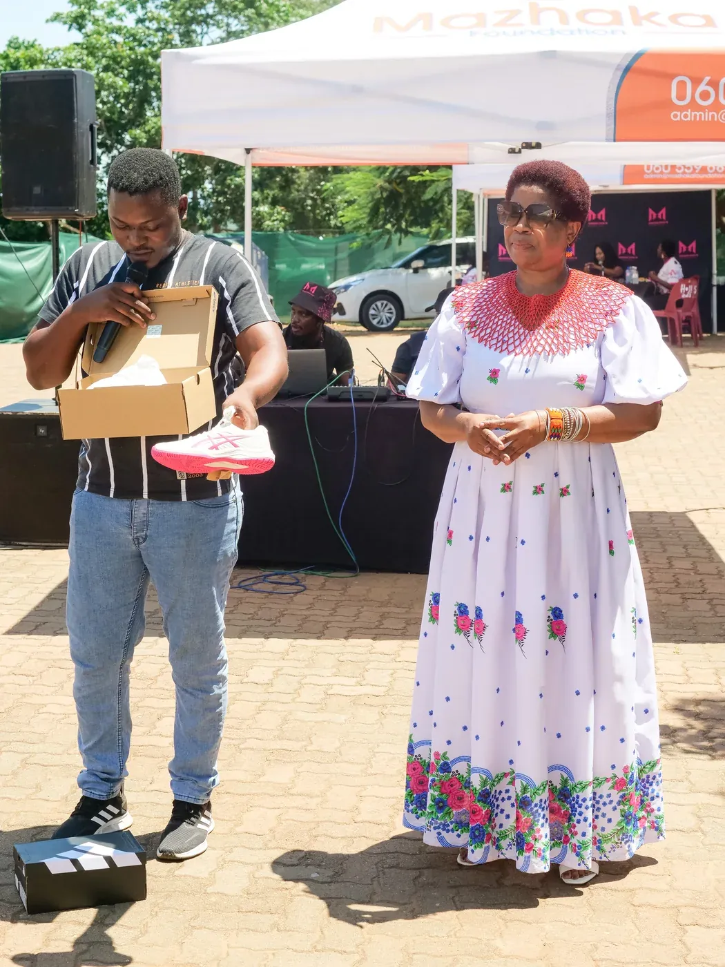 A man holding a pair of school shoes beside a woman in traditional dress at a school-shoe donation event